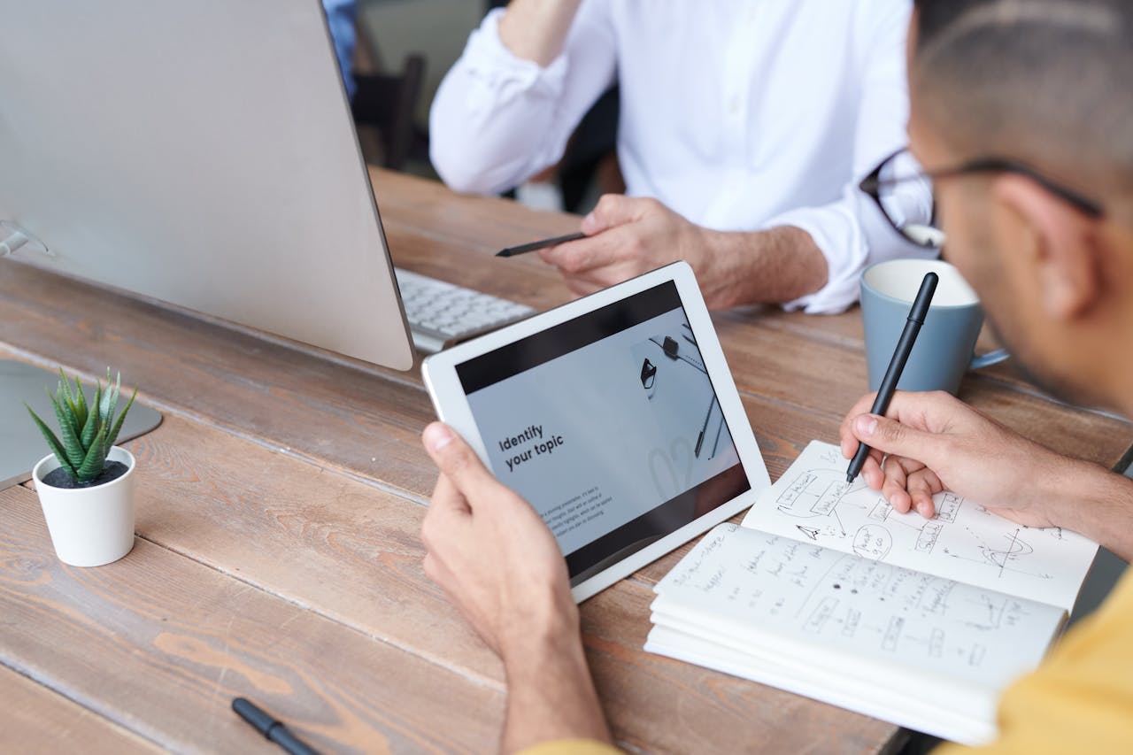 Two professionals collaborating using a tablet and notebook at a wooden table indoors.